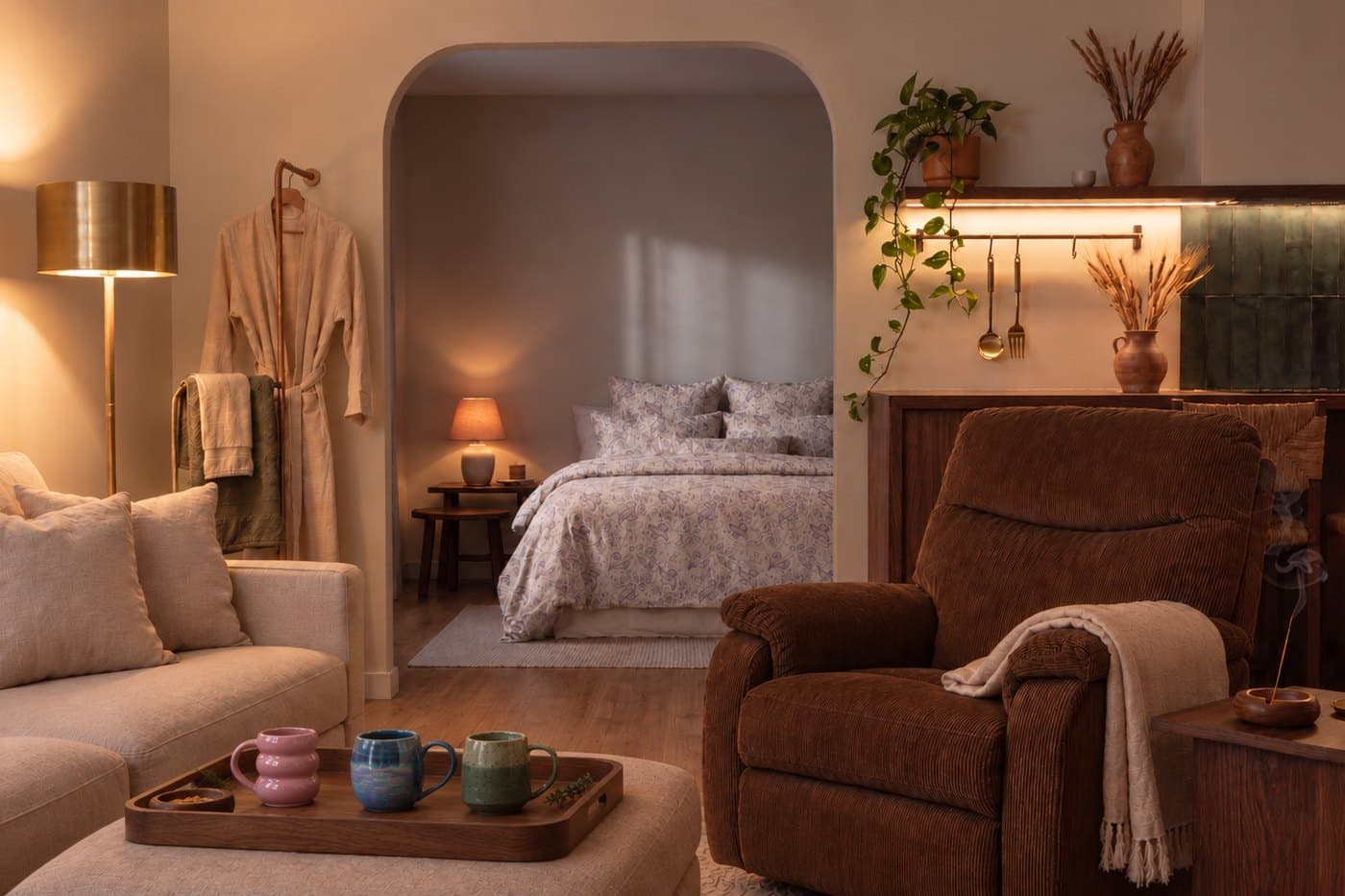 A warmly lit living room with a tray of handmade Karmaura mugs, a robe and towels by the door, and a made bed through an archway